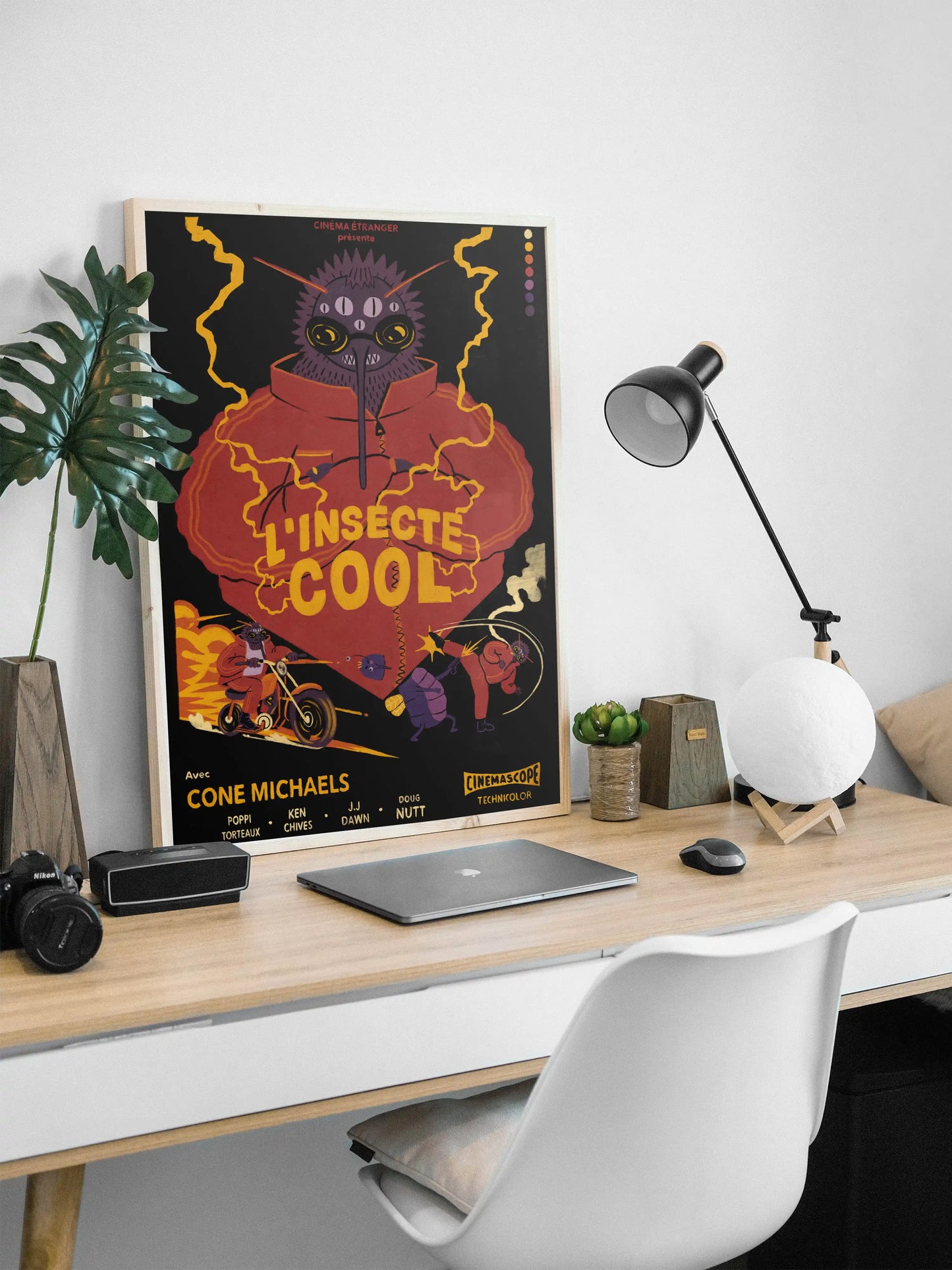 Framed ‘L’insecte Cool’ French bug-noir poster by NYC artist Max Kolo displayed on a modern office desk with laptop, lamp, plants, and camera gear, featuring a bold retro insect character illustration.