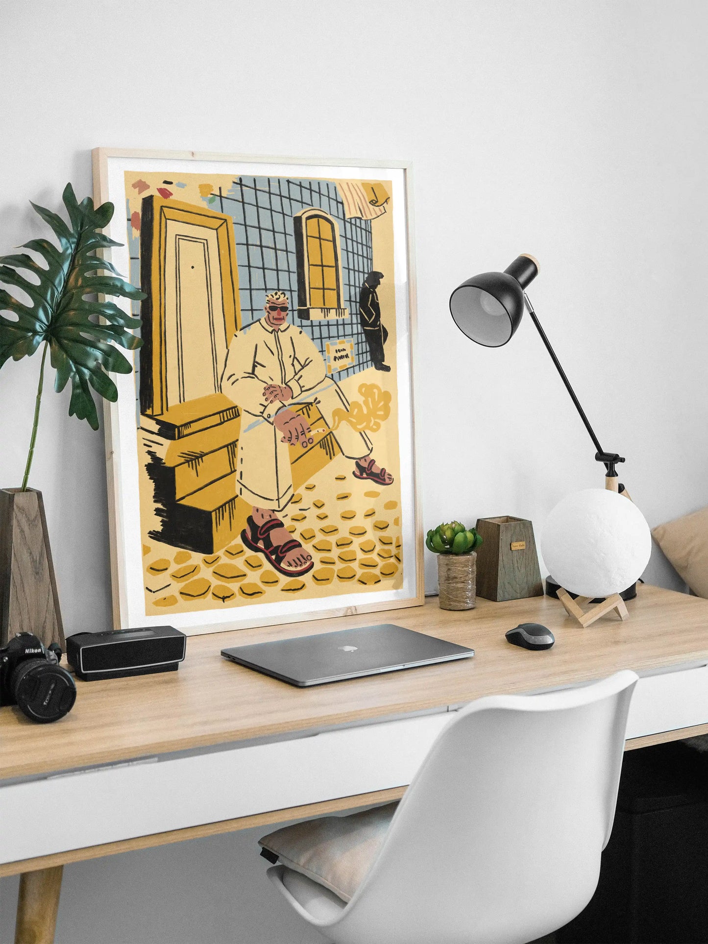 Framed ‘Lisbon’ city illustration poster by NYC artist Max Kolo displayed on a modern office desk with laptop, lamp, plants, and camera gear, featuring a stylized Lisbon man sitting on sunlit tiled steps.