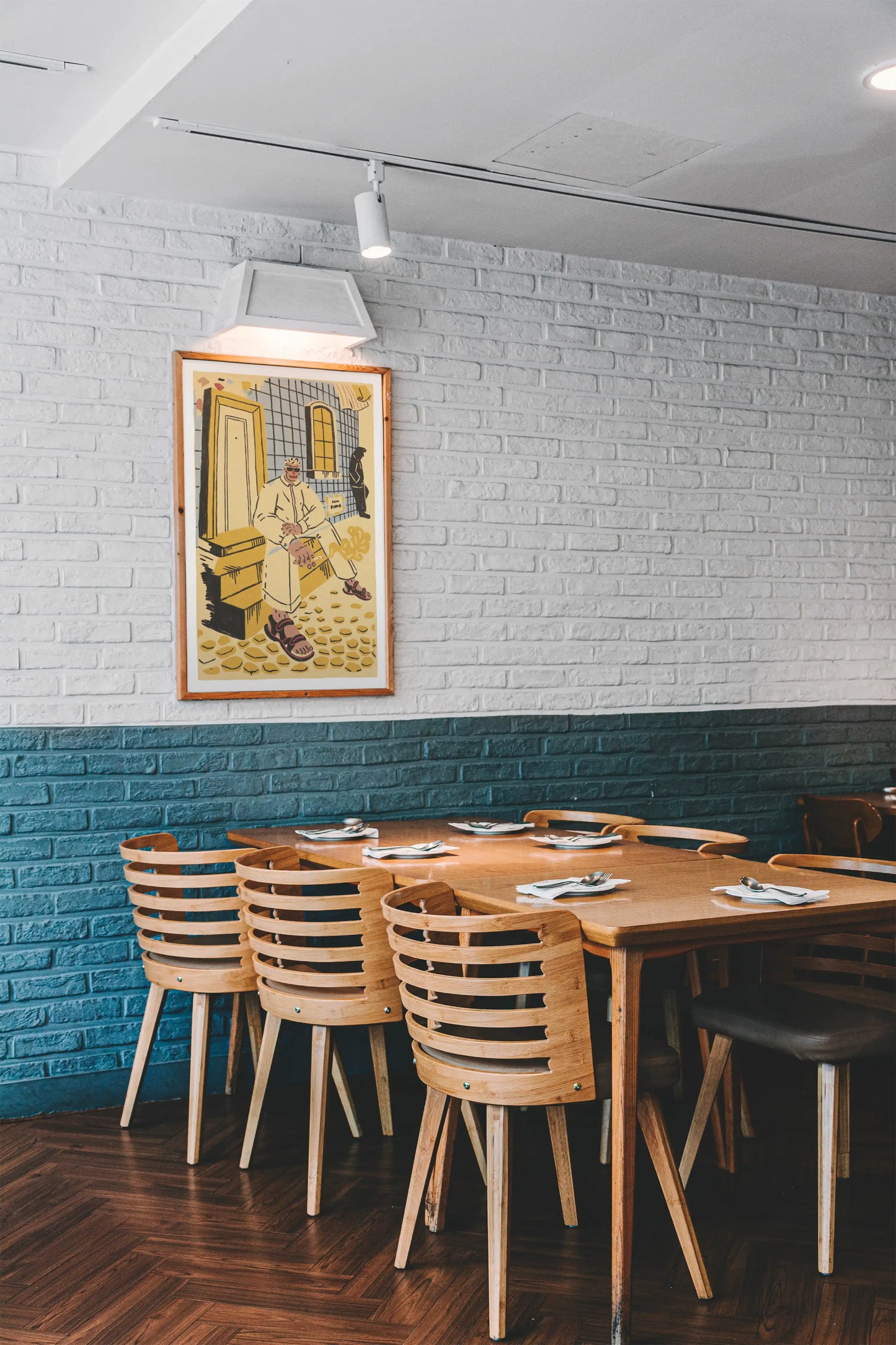 Framed ‘Lisbon’ city illustration poster by NYC artist Max Kolo hanging on a white brick wall in a modern restaurant, featuring a stylized Lisbon man sitting on sunlit tiled steps.