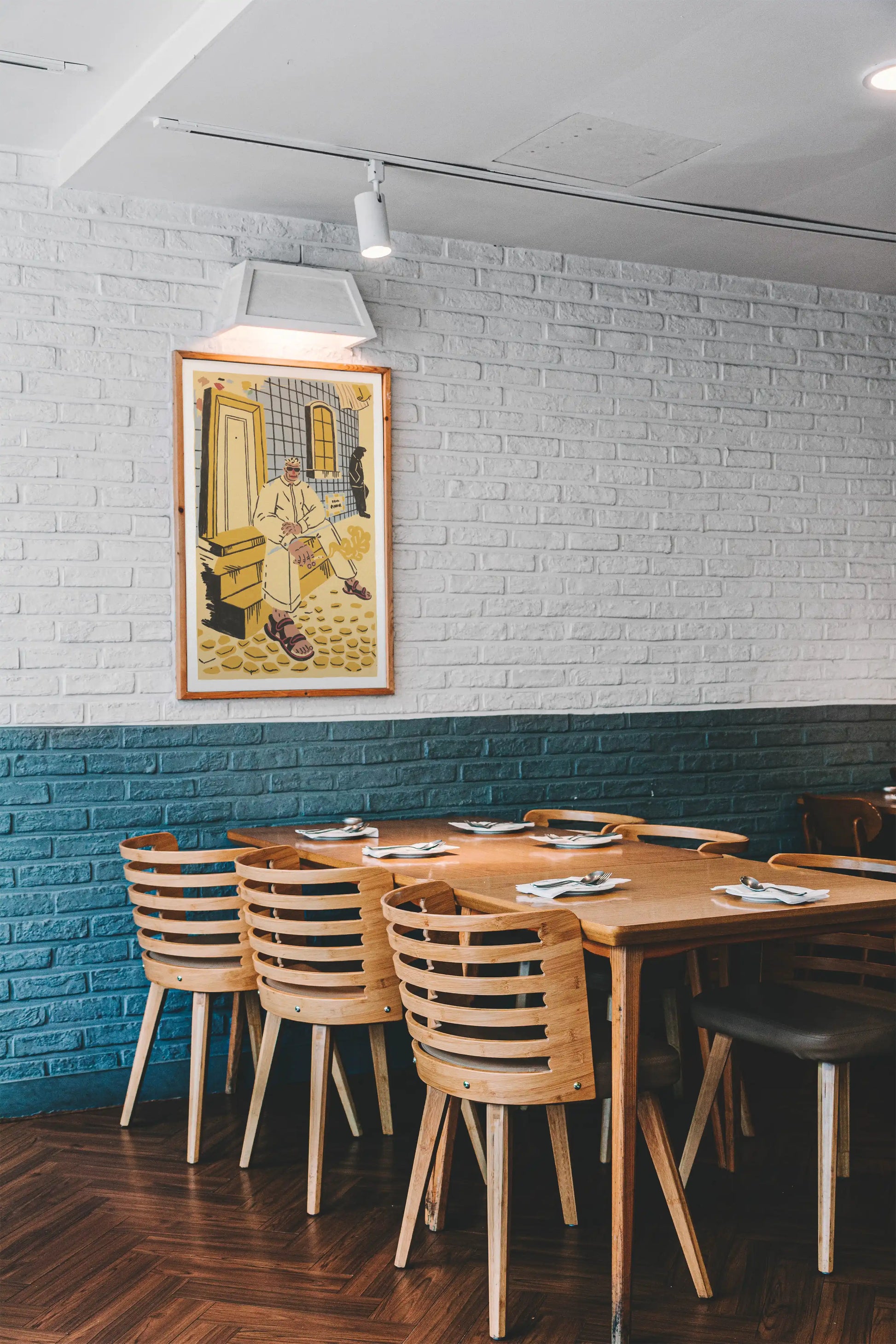 Framed ‘Lisbon’ city illustration poster by NYC artist Max Kolo hanging on a white brick wall in a modern restaurant, featuring a stylized Lisbon man sitting on sunlit tiled steps.