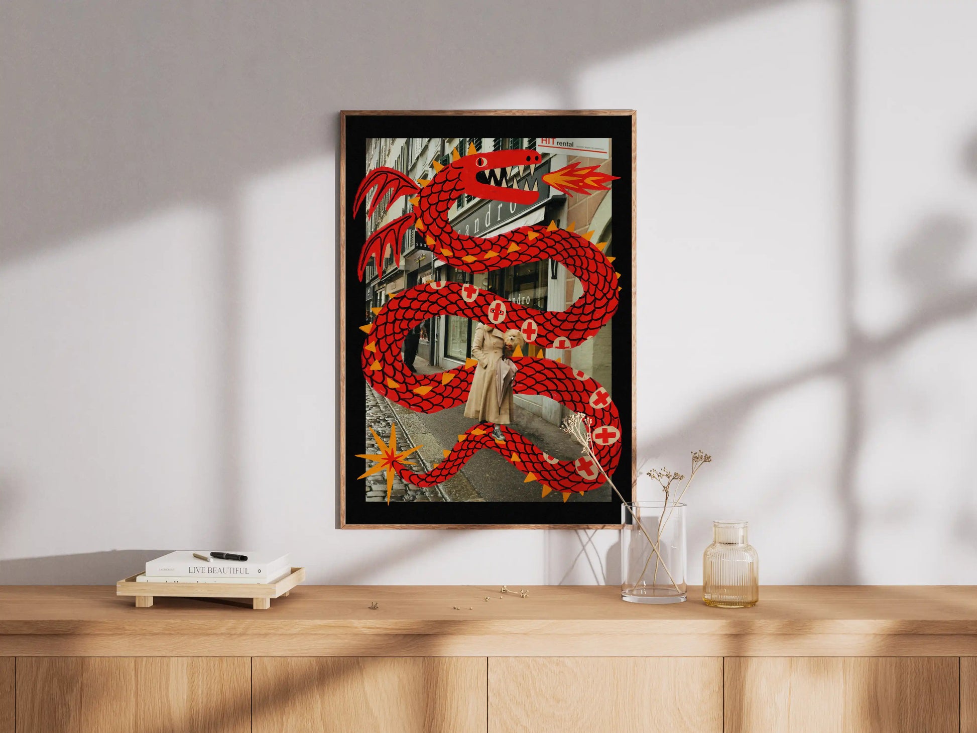 Framed artwork of a Lucerne street scene with a woman, her dog, and a large illustrated red dragon. By  NYC Artist Max Kolo on a wooden surface with a white wall background