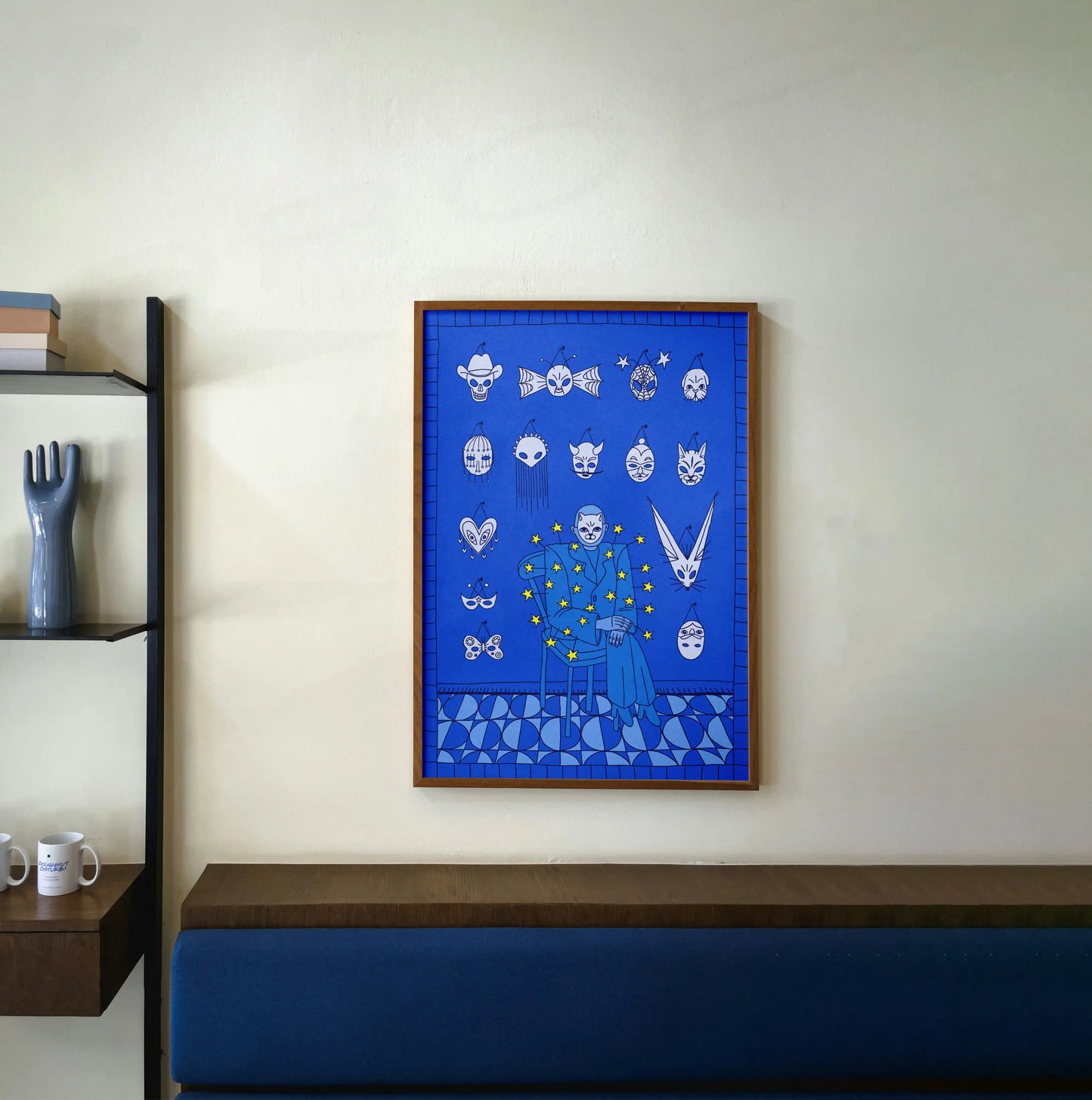 Framed Mask Salesman art print by Max Kolo hanging on a light wall above a wooden bench, showcasing a surreal blue design with expressive white masks.