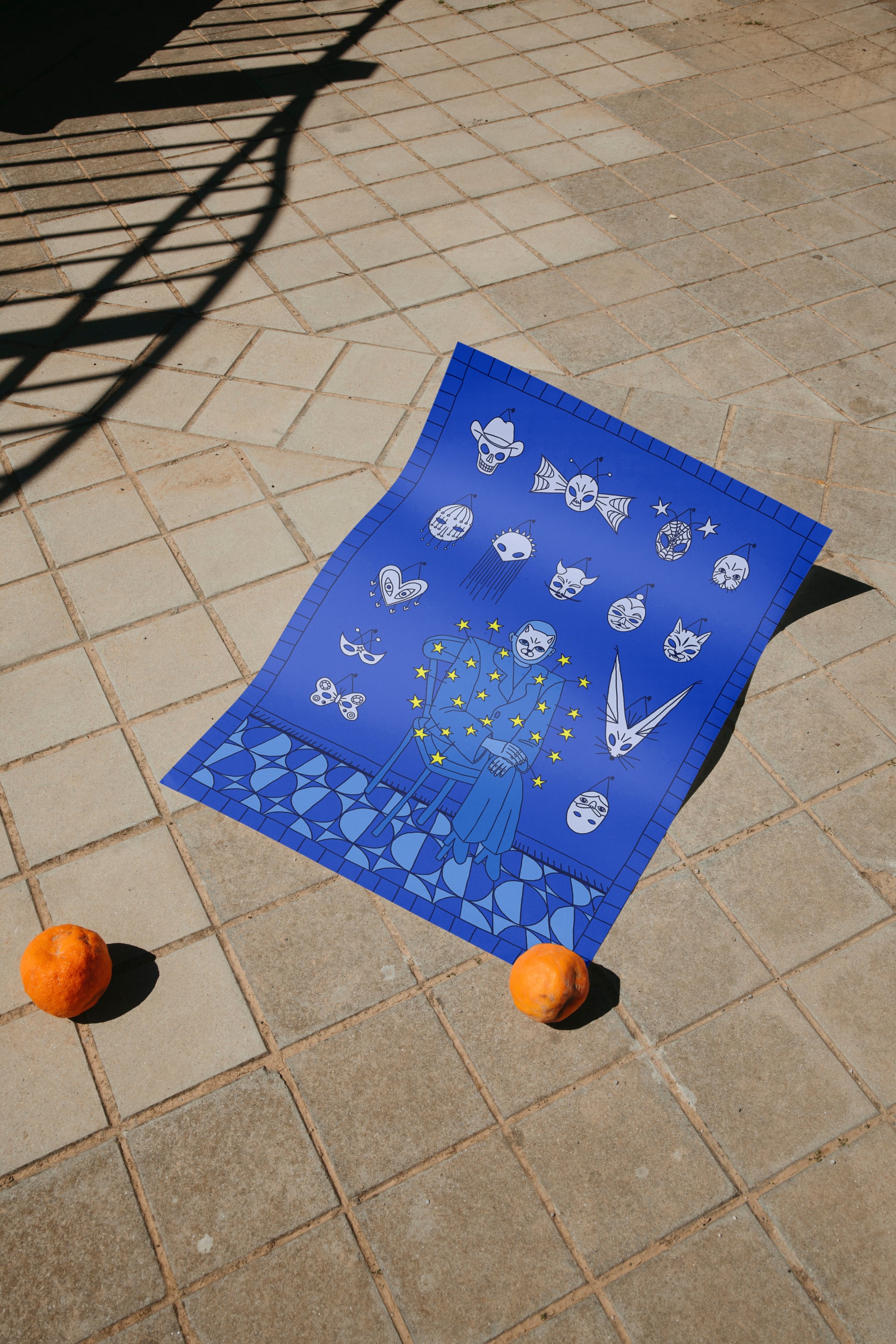 Blue Mask Salesman art print by NYC artist Max Kolo laid on outdoor stone pavement with two oranges, featuring surreal white mask illustrations and abstract shapes.