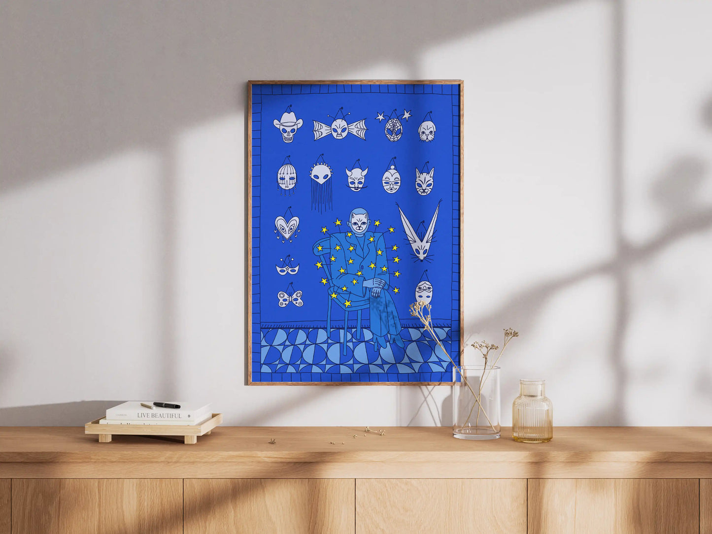 Framed Mask Salesman art print by Max Kolo hanging on a white wall above a wooden credenza, featuring a surreal blue design with expressive white masks.