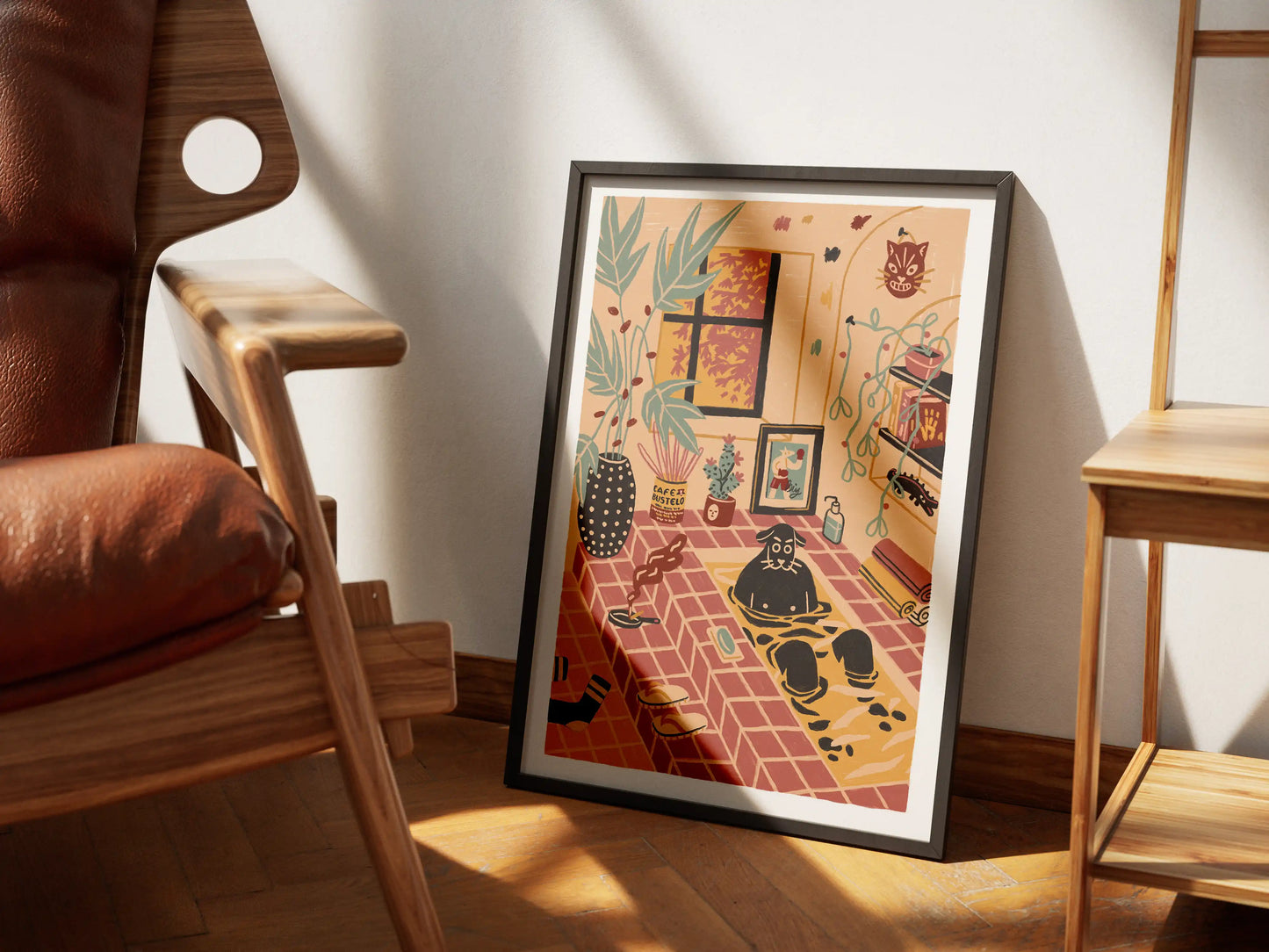 Framed ‘Me Time’ art print by Max Kolo featuring a cozy interior with a dog soaking in a warm tiled bath surrounded by plants and whimsical décor, displayed on a wooden floor beside a mid-century leather chair and a wooden ladder shelf in soft natural light.