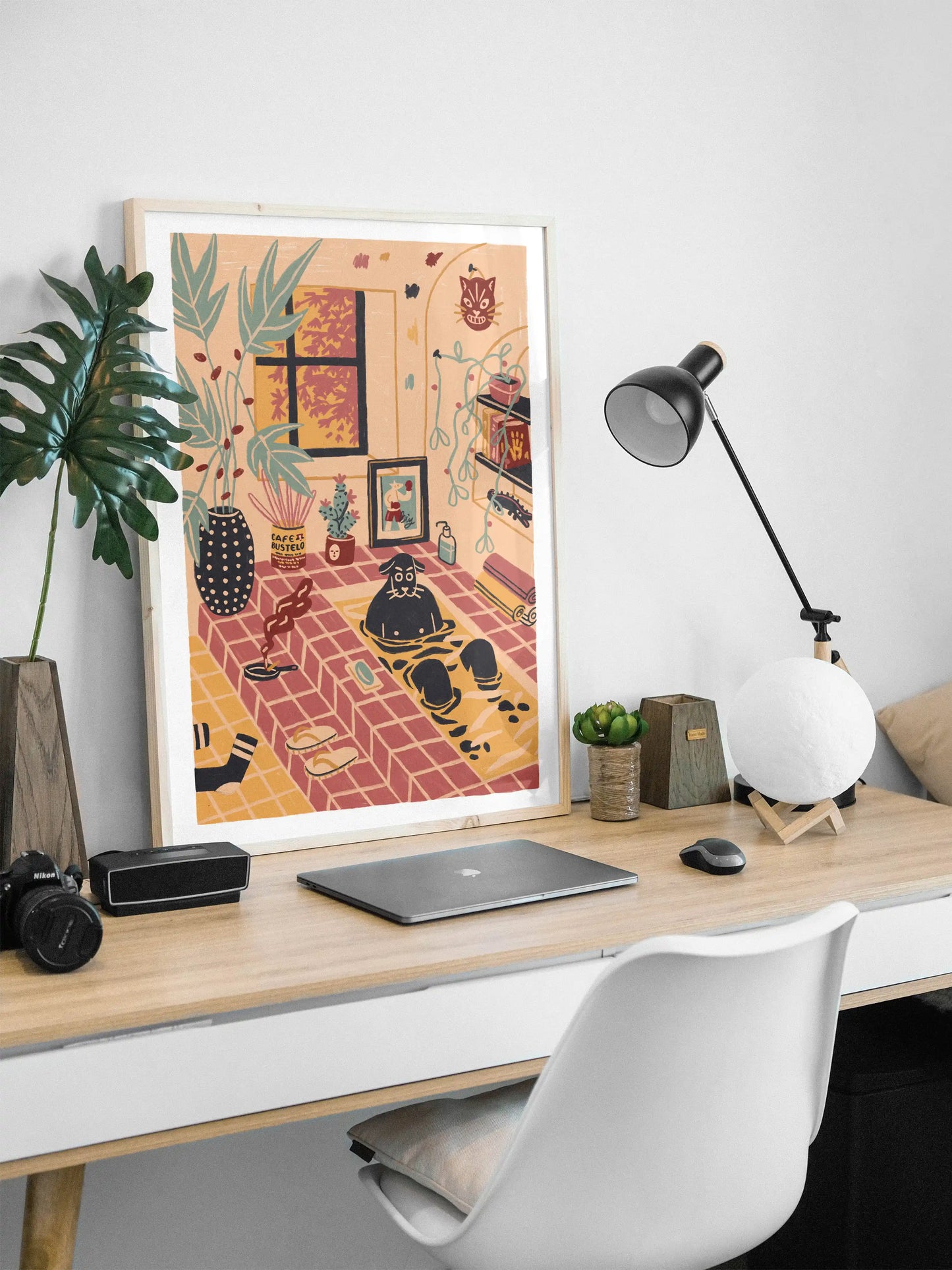 Framed ‘Me Time’ art print by Max Kolo displayed on a modern wood desk setup. The illustration shows a cozy indoor room with a black dog relaxing in a tiled bath surrounded by plants, artwork, and warm décor. A laptop, desk lamp, plant, and white chair sit in the foreground against a clean white wall.
