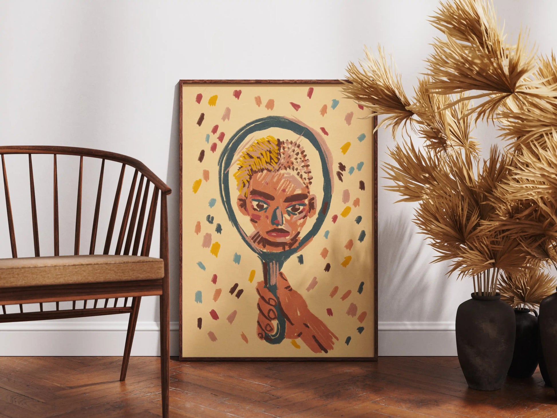 Framed "Mirror" art print by Max Kolo on a wooden floor beside a wooden chair and decorative dried plants, featuring a stylized painted face reflected in a hand-held mirror.
