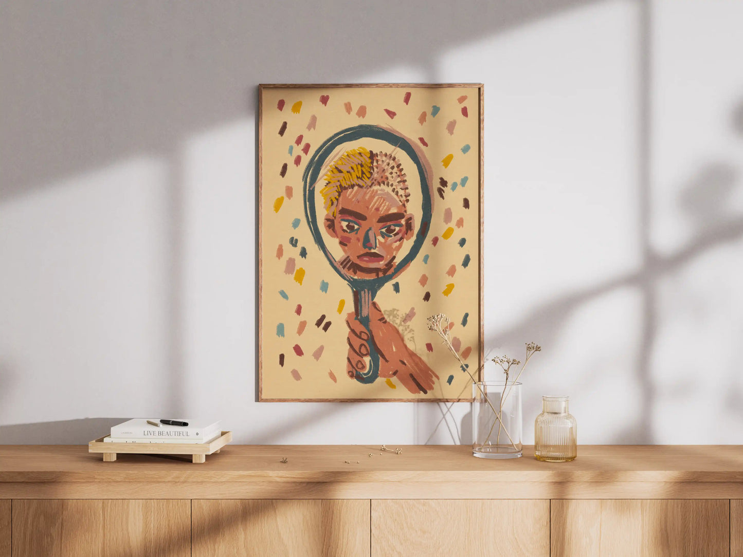 Framed "Mirror" art print by NYC artist Max Kolo hanging on a white wall above a wooden sideboard, featuring a stylized painted face reflected in a hand-held mirror illustration.