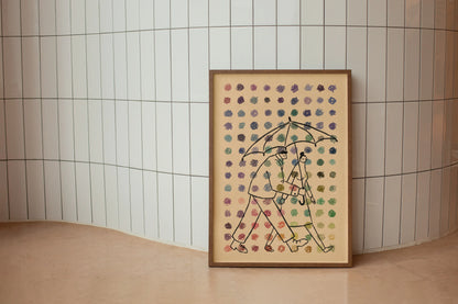 Framed artwork of two people under an umbrella with a patterned background and dots of multiple colors in the foreground set against a tiled wall. Artwork by New York City Artist Max Kolo.