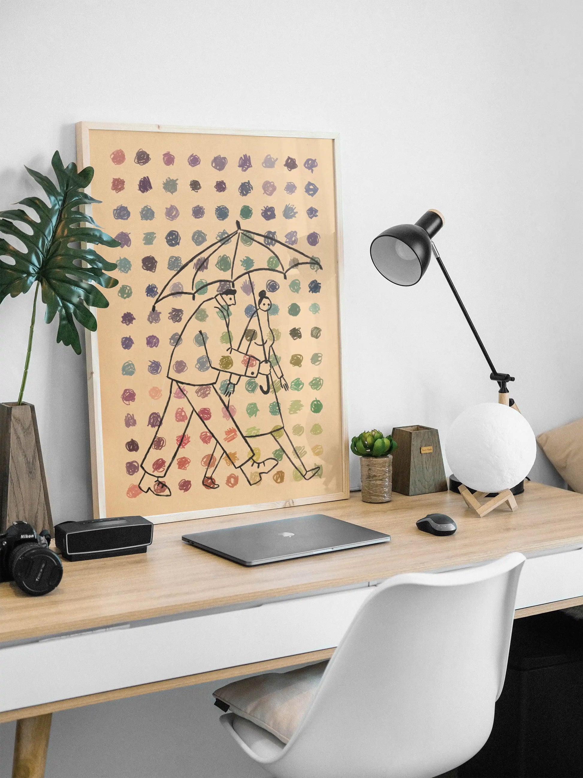 Framed illustration by NYC artist Max Kolo showing two stylized figures walking under a single umbrella beneath colorful rain, displayed on a modern home desk setup with a wooden tabletop, office lamp, laptop, small plants, and a white chair against a clean white wall.