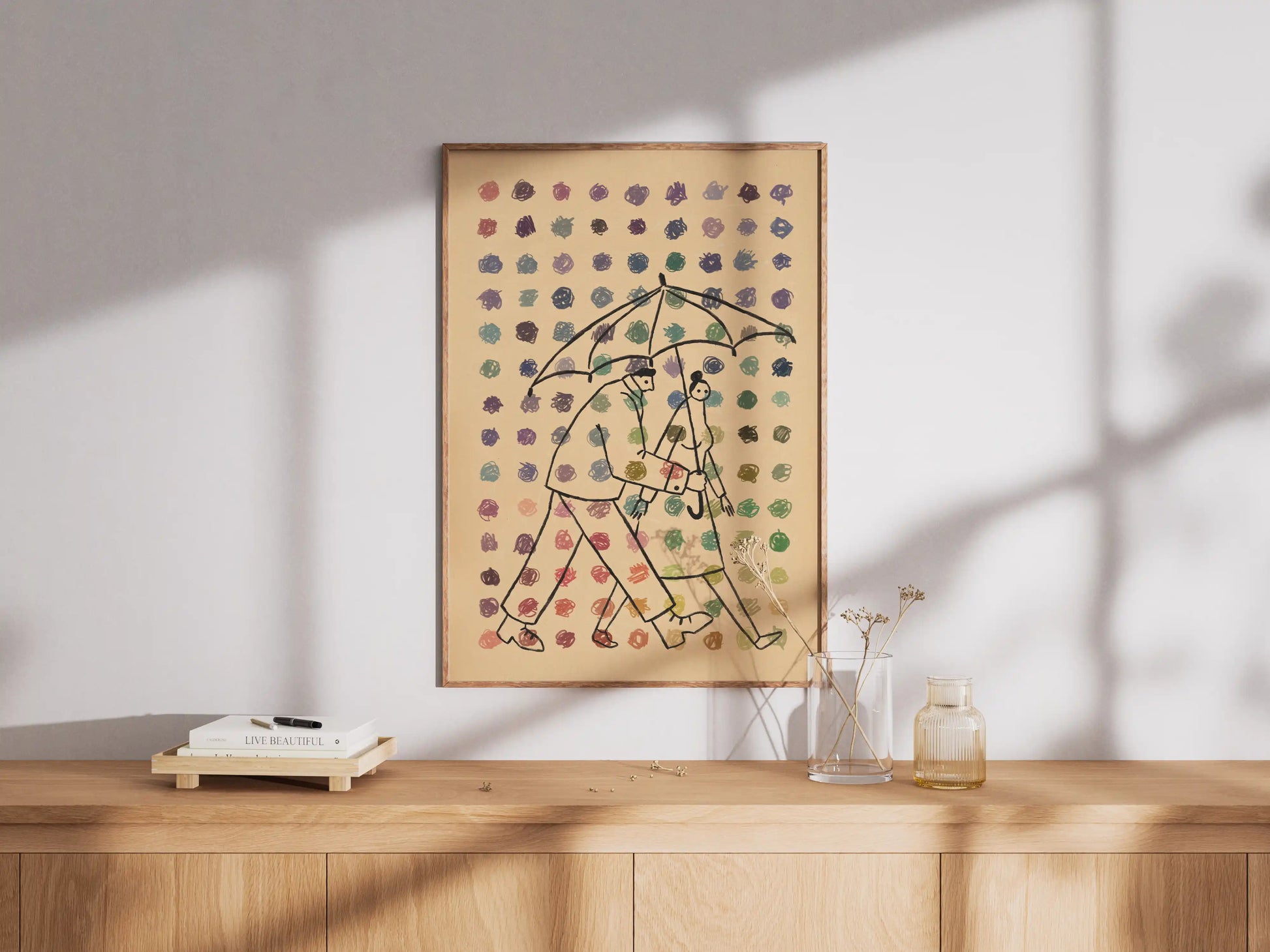 Framed illustration by NYC artist Max Kolo featuring two stylized figures walking together under a single umbrella as colorful raindrops fall against a beige background, displayed on a wooden sideboard in soft natural light.