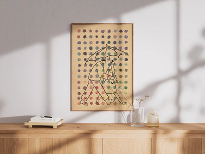 Framed illustration by NYC artist Max Kolo featuring two stylized figures walking together under a single umbrella as colorful raindrops fall against a beige background, displayed on a wooden sideboard in soft natural light.
